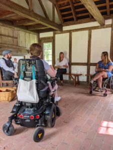 Four people, one in a mechanical wheelchair and one in a powerchair, conversing while seated in a room of a historic building with wood beams.