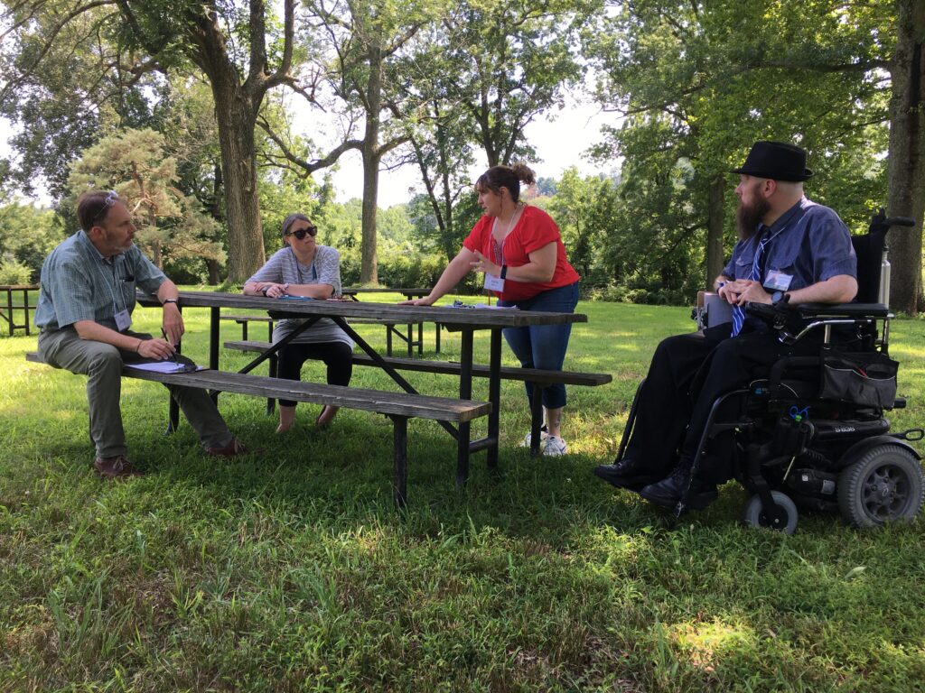Four people gather around a picnic table in a grassy area surrounded by trees--two of them are seated, one is leaning on the table while standing and one is slightly off to the right in a powerchair.