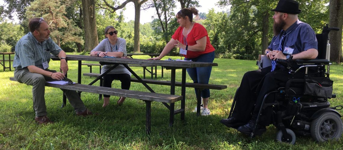Four people gather around a picnic table in a grassy area surrounded by trees--two of them are seated, one is leaning on the table while standing and one is slightly off to the right in a powerchair.